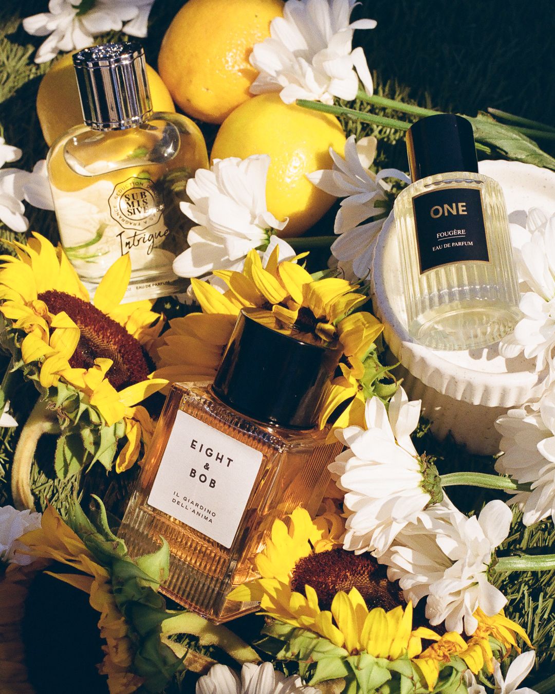 Luxury perfume bottles including Eight & Bob and ONE eau de parfum arranged with lemons and white and yellow flowers.
