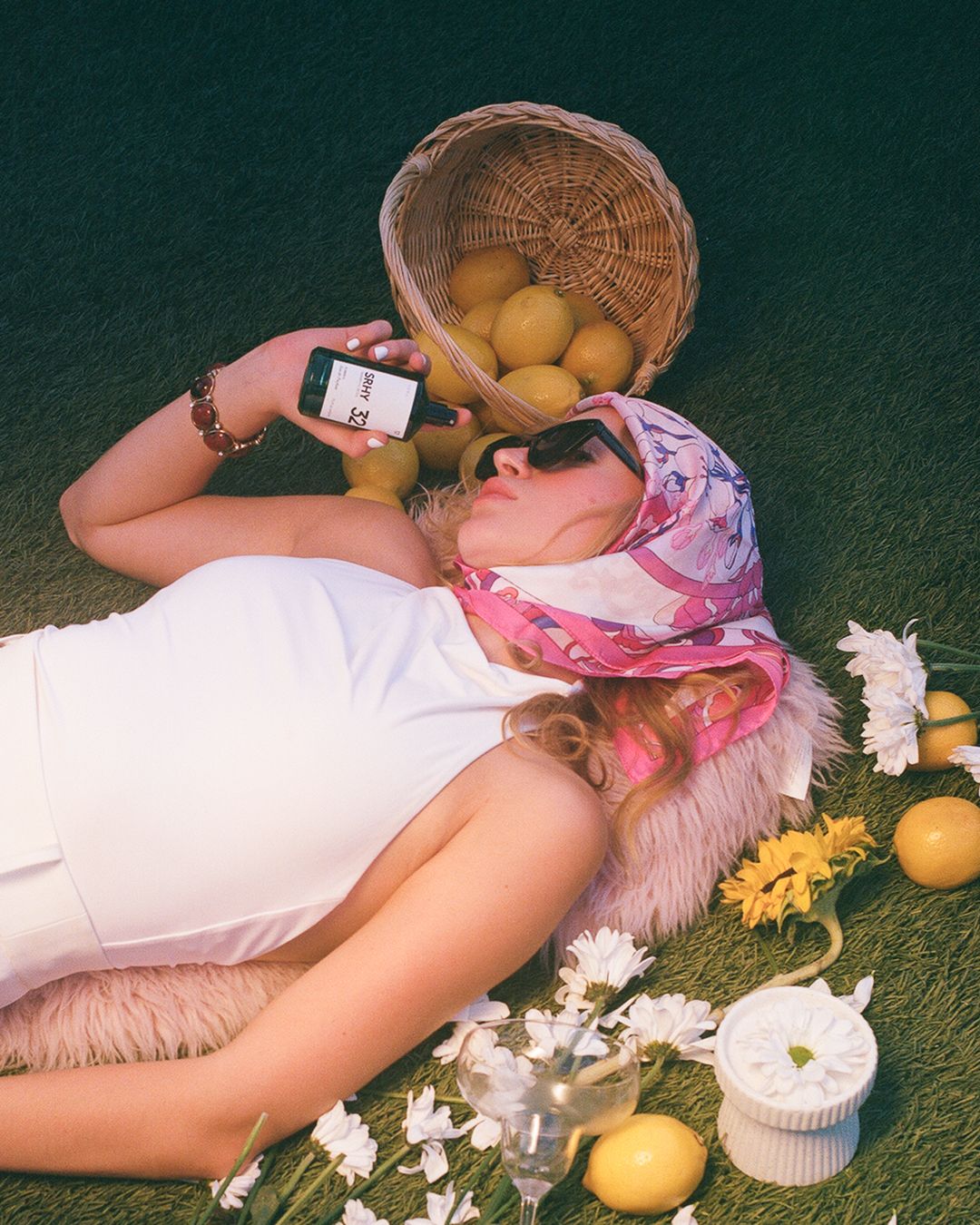 Woman lying on grass surrounded by lemons and flowers, holding a phone, with a wicker basket overhead.