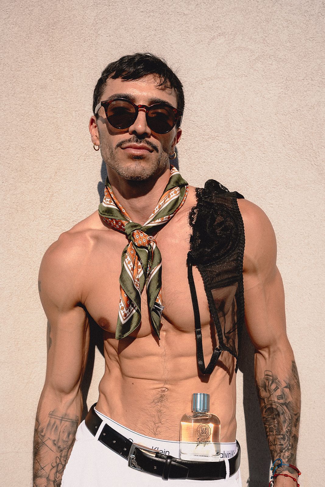 Shirtless tattooed man wearing sunscarf and sunglasses holding Eight and Oak perfume bottle outdoors.