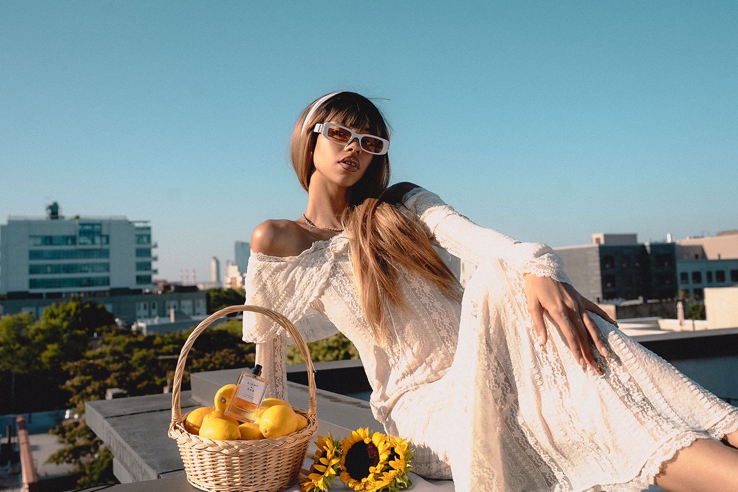 Stylish woman in white lace dress holding basket of lemons with sunflowers on rooftop terrace.
