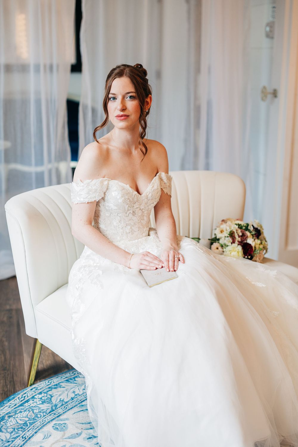 Elegant off-shoulder white wedding gown with lace details and floral bouquet on cream armchair against sheer curtains.