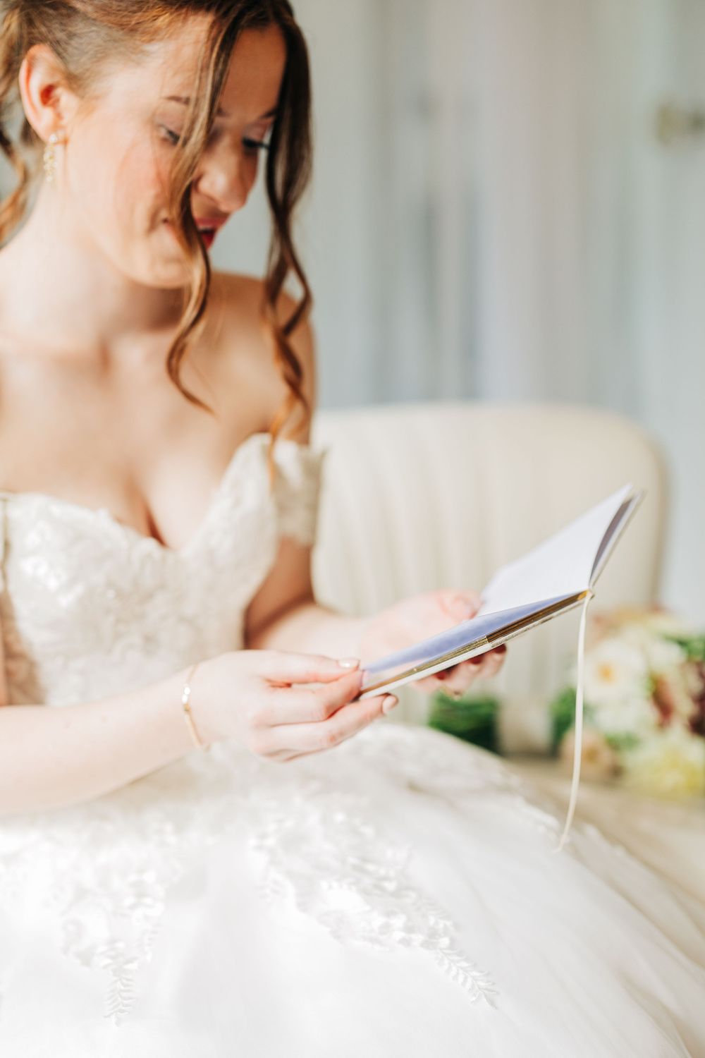 A bride in a white wedding dress sits at a table writing with a pen on paper in a soft, romantic setting.