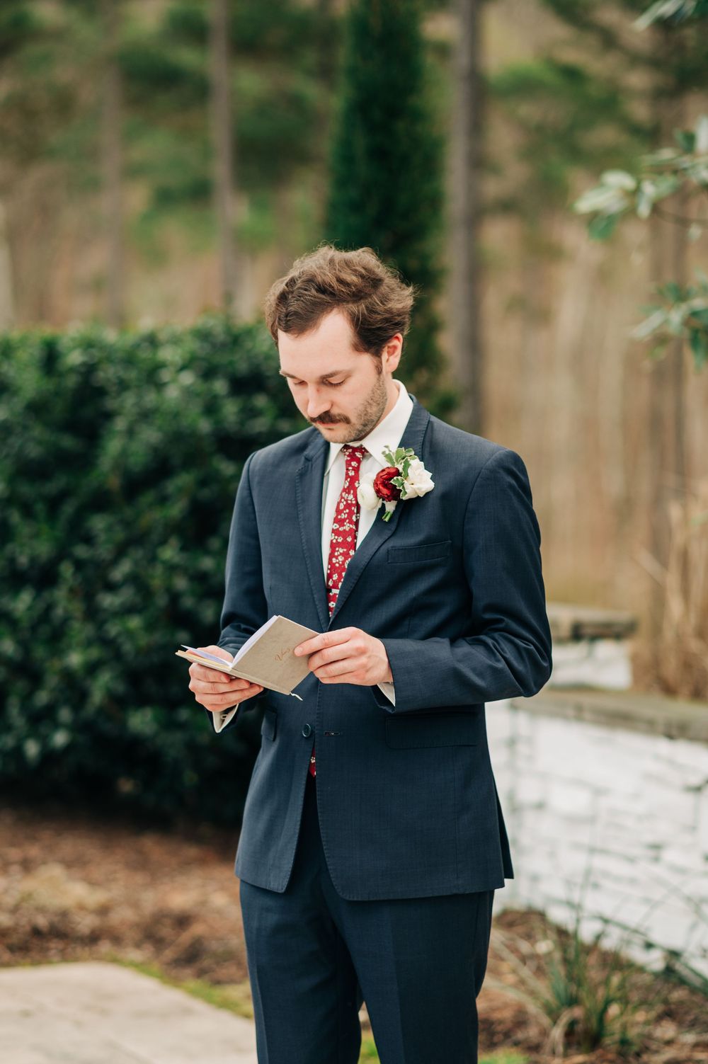 A person in a dark suit with a red tie reads from a paper in an outdoor winter setting.