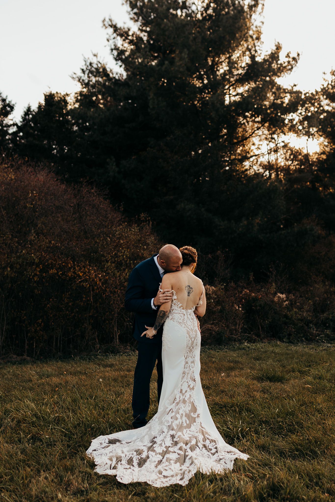 A couple in wedding attire embraces in a scenic field at sunset with trees in the background.