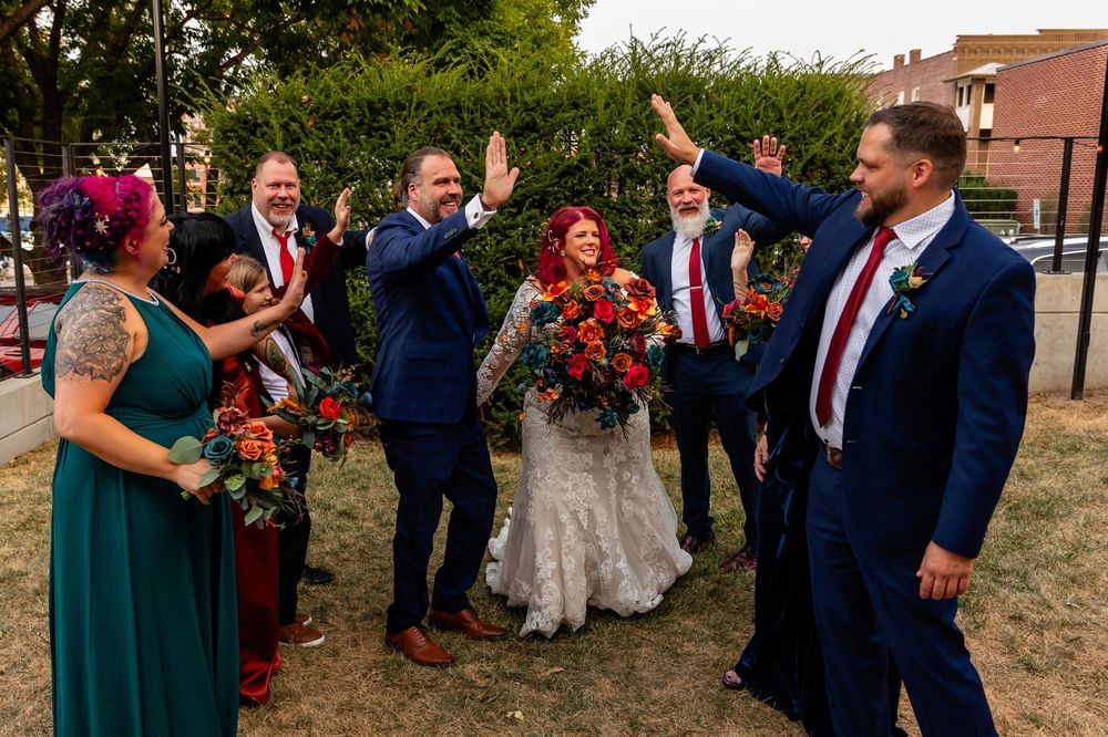 Wedding party in navy suits and teal gowns celebrating with high fives around bride holding vibrant red floral bouquet at Founders One Nine