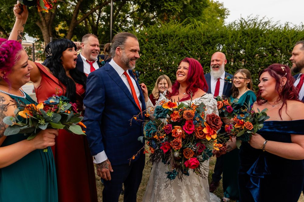 Bride with red hair holding colorful bouquet stands with groom in blue suit surrounded by cheering wedding party at Founders One Nine