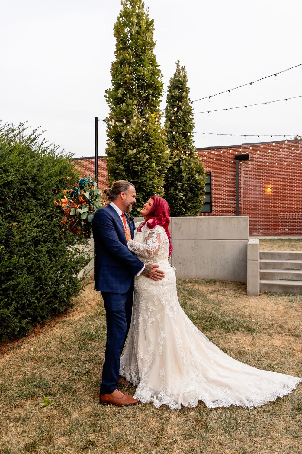 Bride in lace gown and groom in navy suit share romantic moment outdoors near cypress trees with string lights at Founders One Nine in Omaha