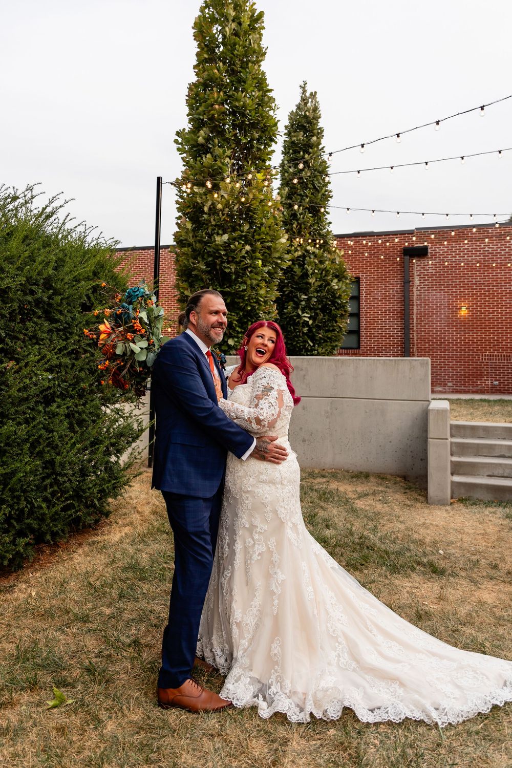 A bride in lace gown with red hair embraces her groom in navy suit outdoors at Founders One Nine in Omaha, NE.