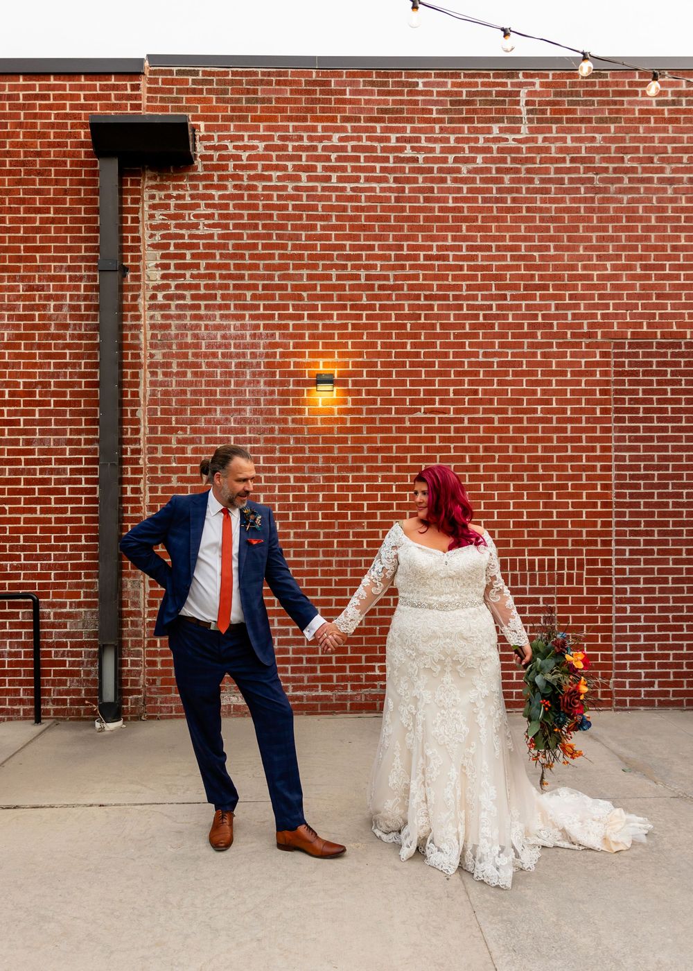 Bride in lace gown with red hair holds hands with groom in navy suit against rustic brick wall outdoors at Founders One Nine in Omaha, NE.