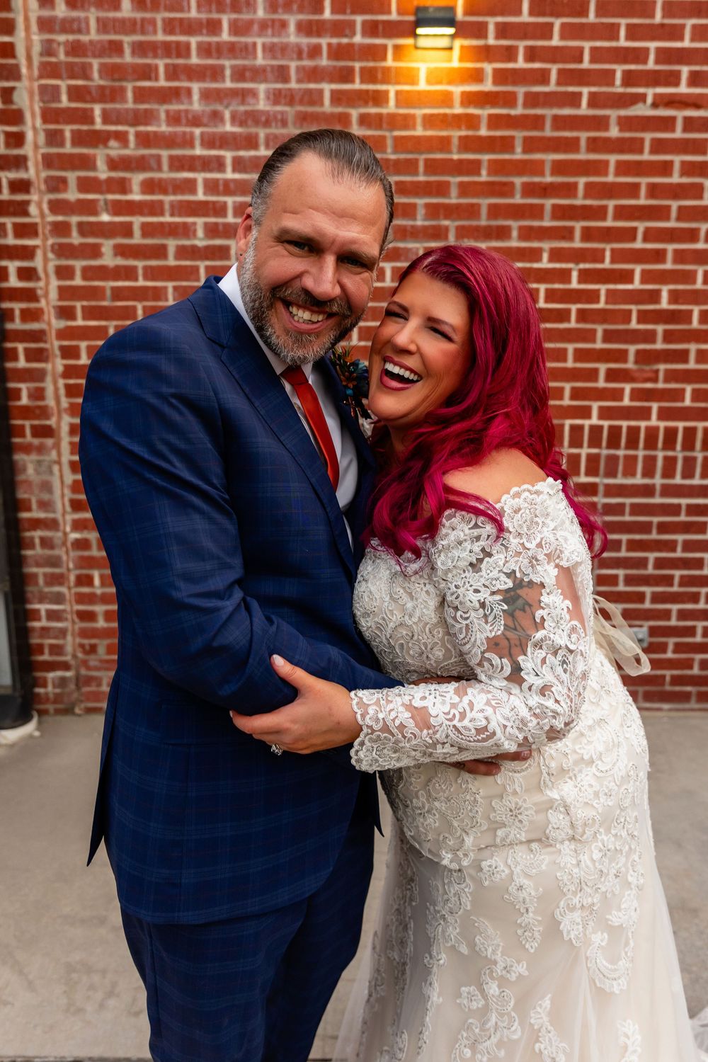 Happy couple embracing in wedding attire, bride in lace gown with red hair, groom in navy suit, brick wall backdrop at Founders One Nine