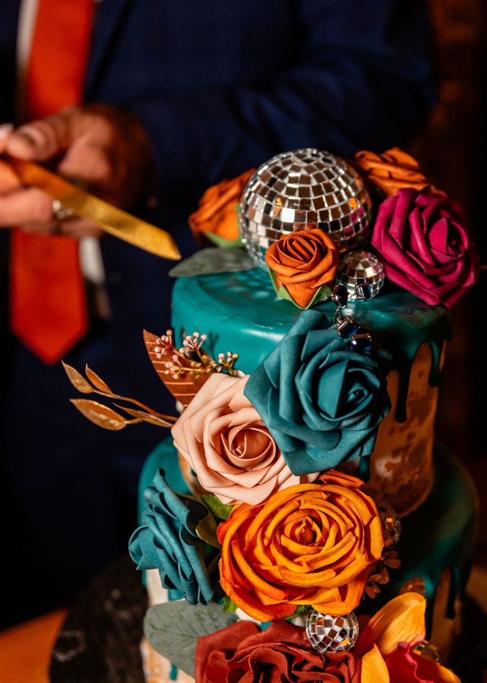 Elegant multi-tier teal wedding cake adorned with colorful roses and disco ball decorations being cut.
