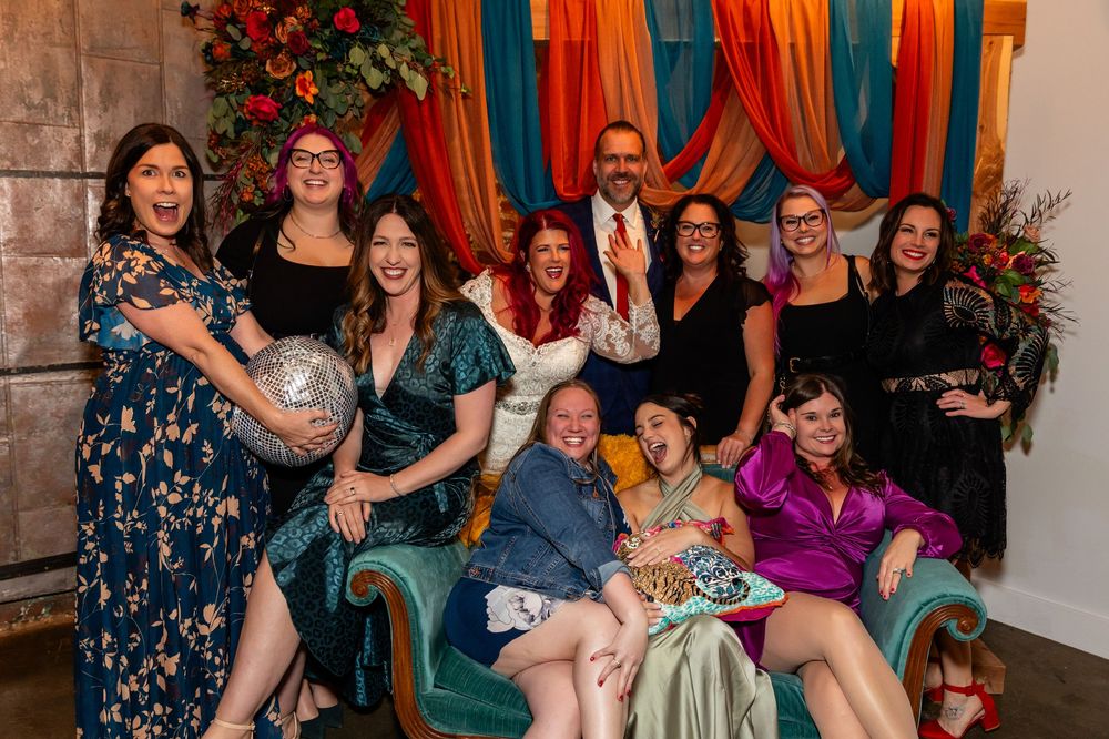 A joyful group of people posing together at a colorful wedding, smiling and laughing in festive attire at Founders One Nine in Omaha, NE