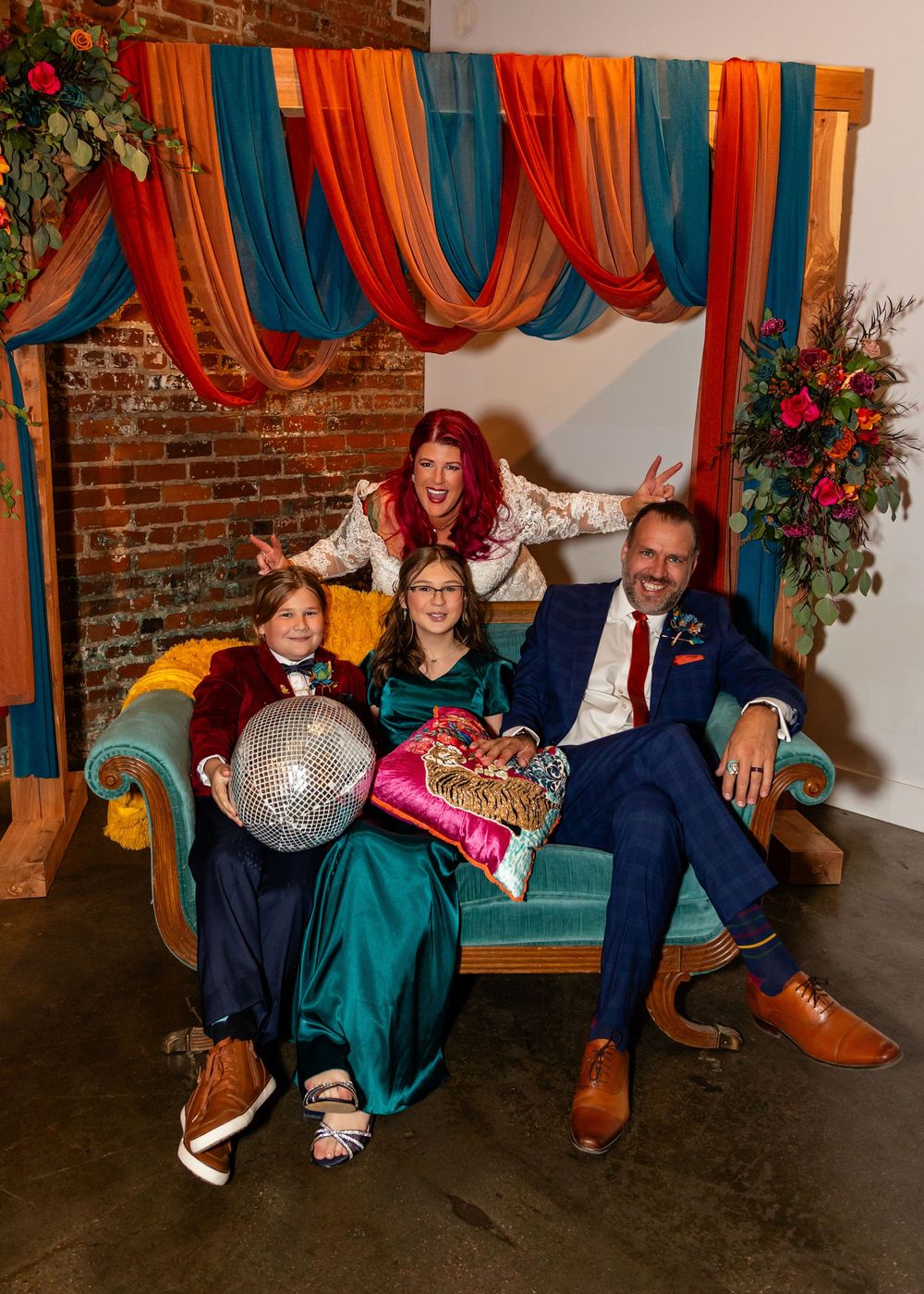 Wedding family portrait on teal velvet sofa under colorful orange and teal draped backdrop with disco ball at Founders One Nine in Omaha, NE