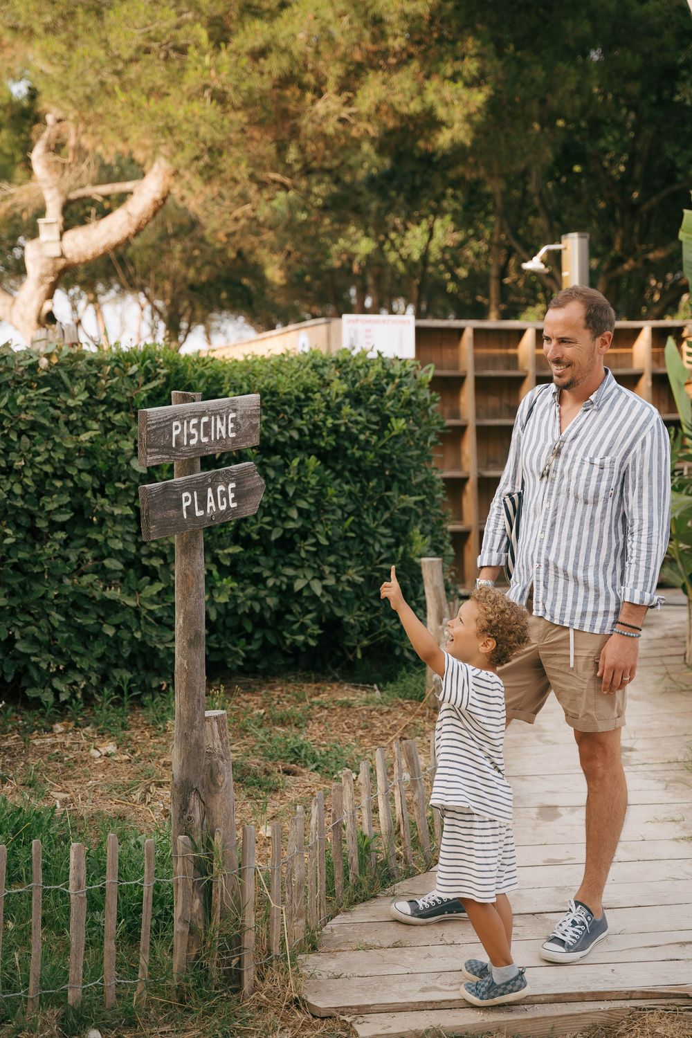 Father and child reading wooden directional signs for Piscine and Plage on a boardwalk path surrounded by greenery.