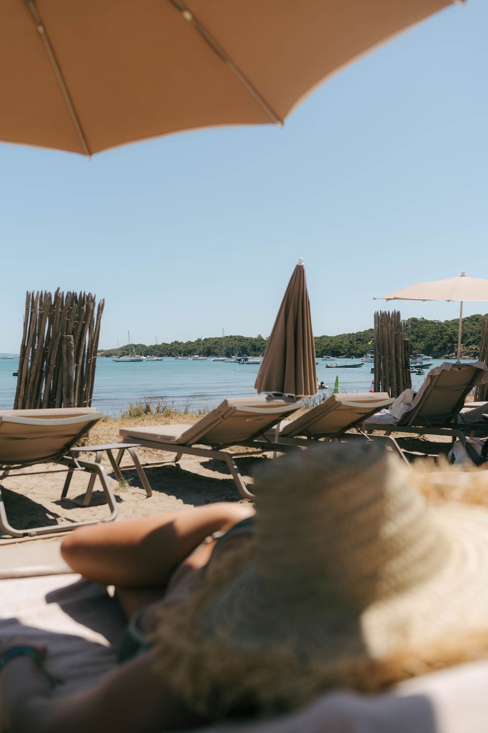 Person relaxing on a beach lounger under a umbrella with calm blue sea and moored boats in the background.