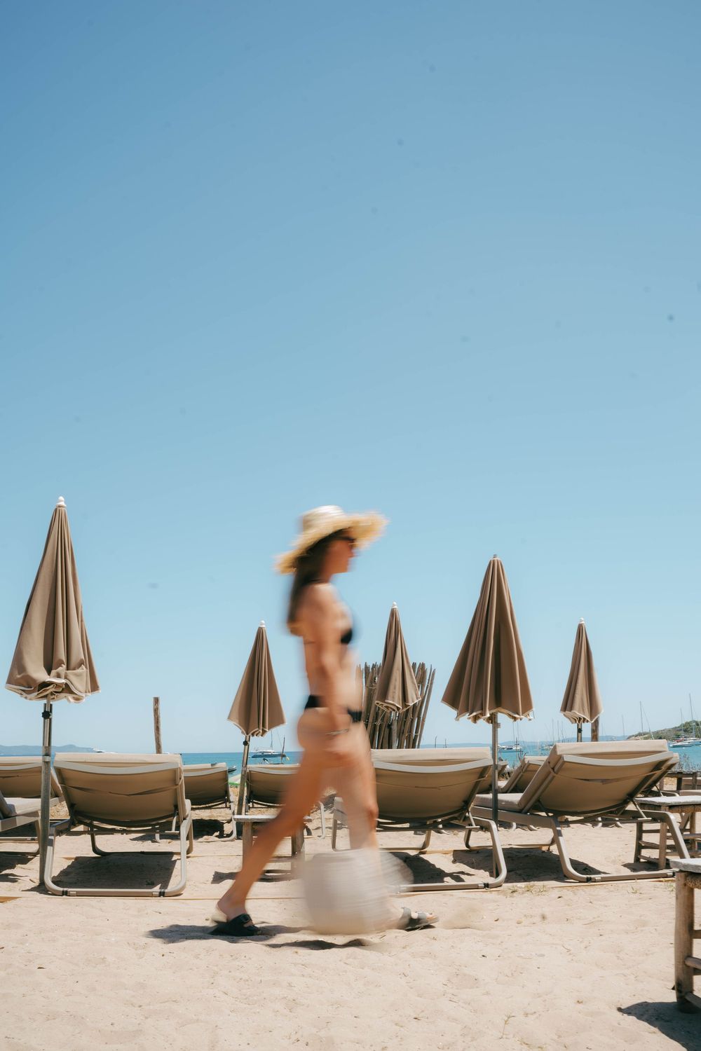 Femme en bikini et chapeau de soleil marchant sur une plage de sable avec des parasols fermés et des chaises longues par une journée ensolei