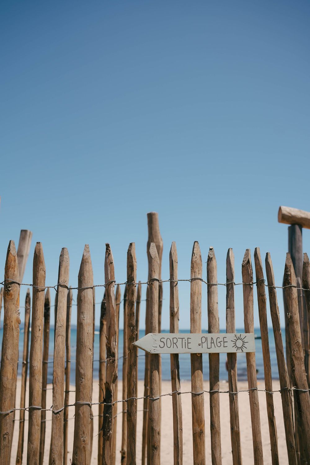 Clôture de plage en bois avec un panneau français indiquant Sortie Plage pointant vers la plage sous un ciel bleu clair.