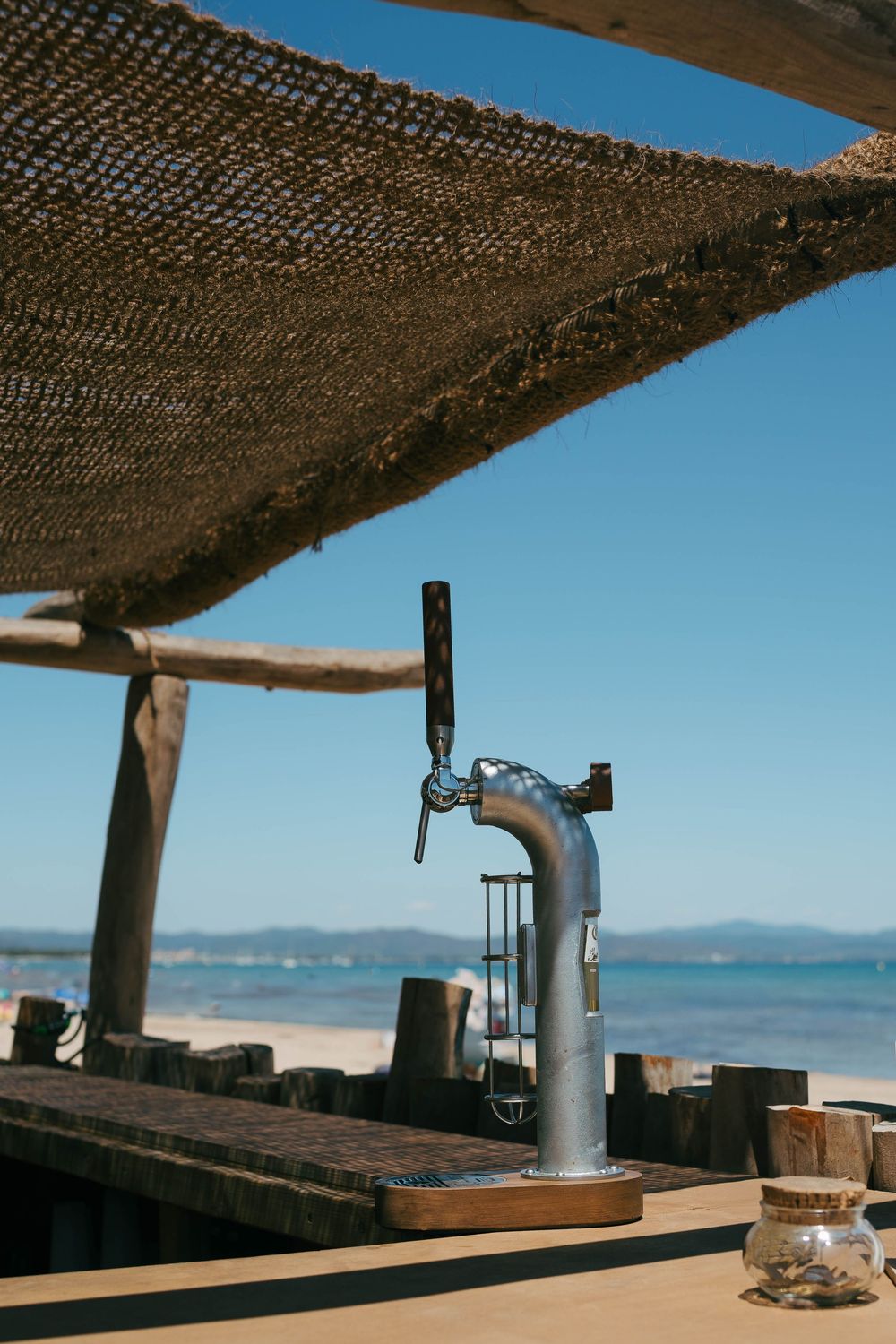 Un robinet de bière argenté dans un bar de plage extérieur avec des filets rustiques au-dessus et des vues sur l'océan sous un ciel bleu cla