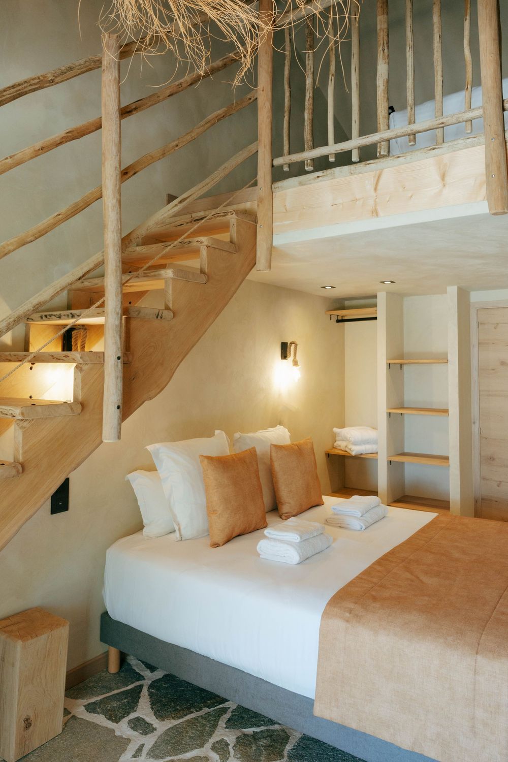 Cozy loft bedroom with wooden staircase, warm beige tones, double bed with orange pillows, and built-in shelving.