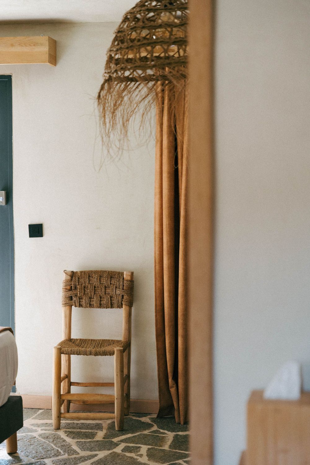 Chaise tissée rustique à côté du rideau en lin et du sol en pierre dans un intérieur méditerranéen minimaliste.