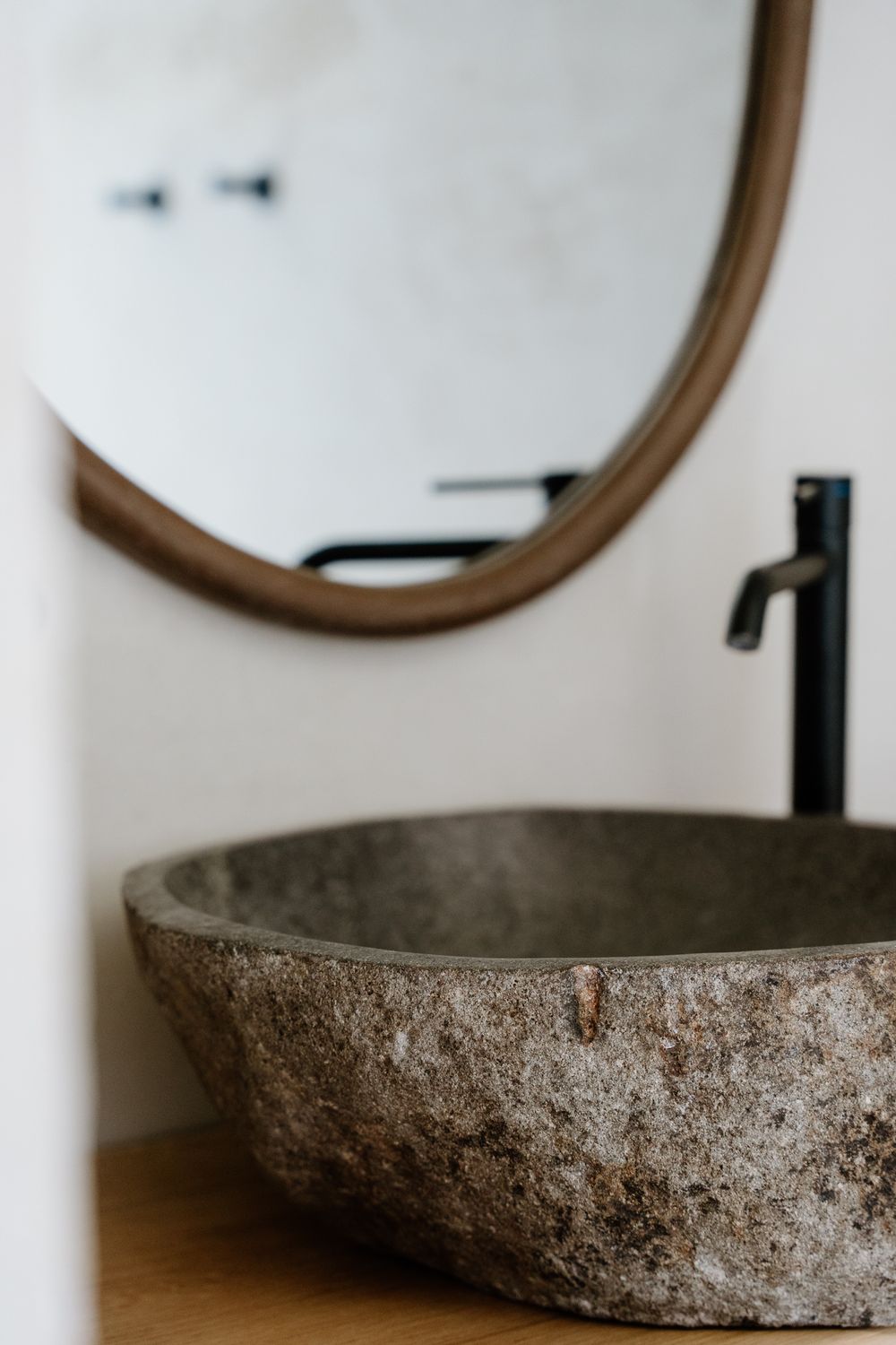 A natural stone vessel sink with black matte faucet on wood vanity, reflected in round wooden framed mirror.