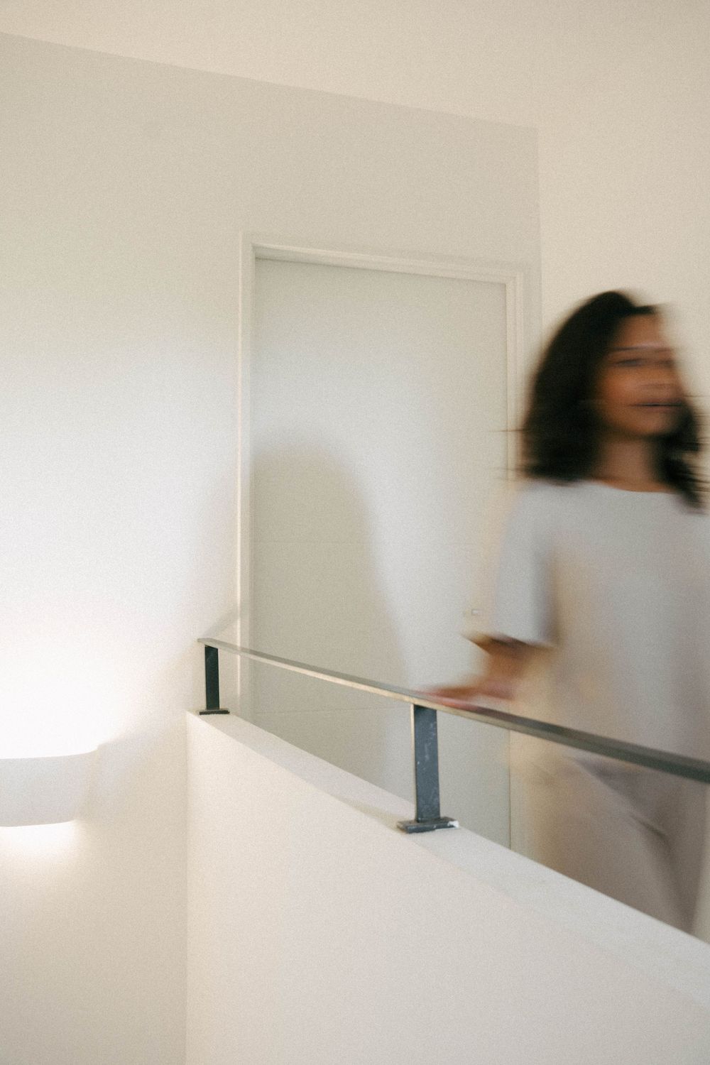Mouvement flou d'une femme marchant devant une rampe d'escalier blanche minimaliste dans un intérieur moderne.