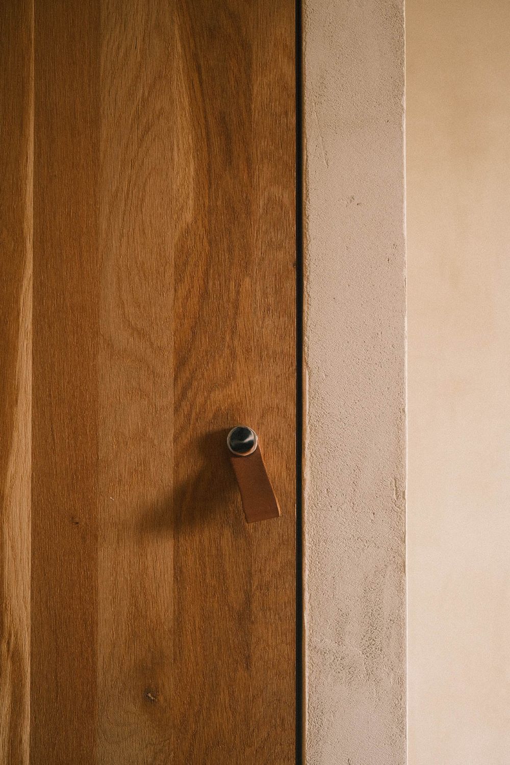 Close-up of a wooden door with a minimalist leather and metal handle against a textured plaster wall.