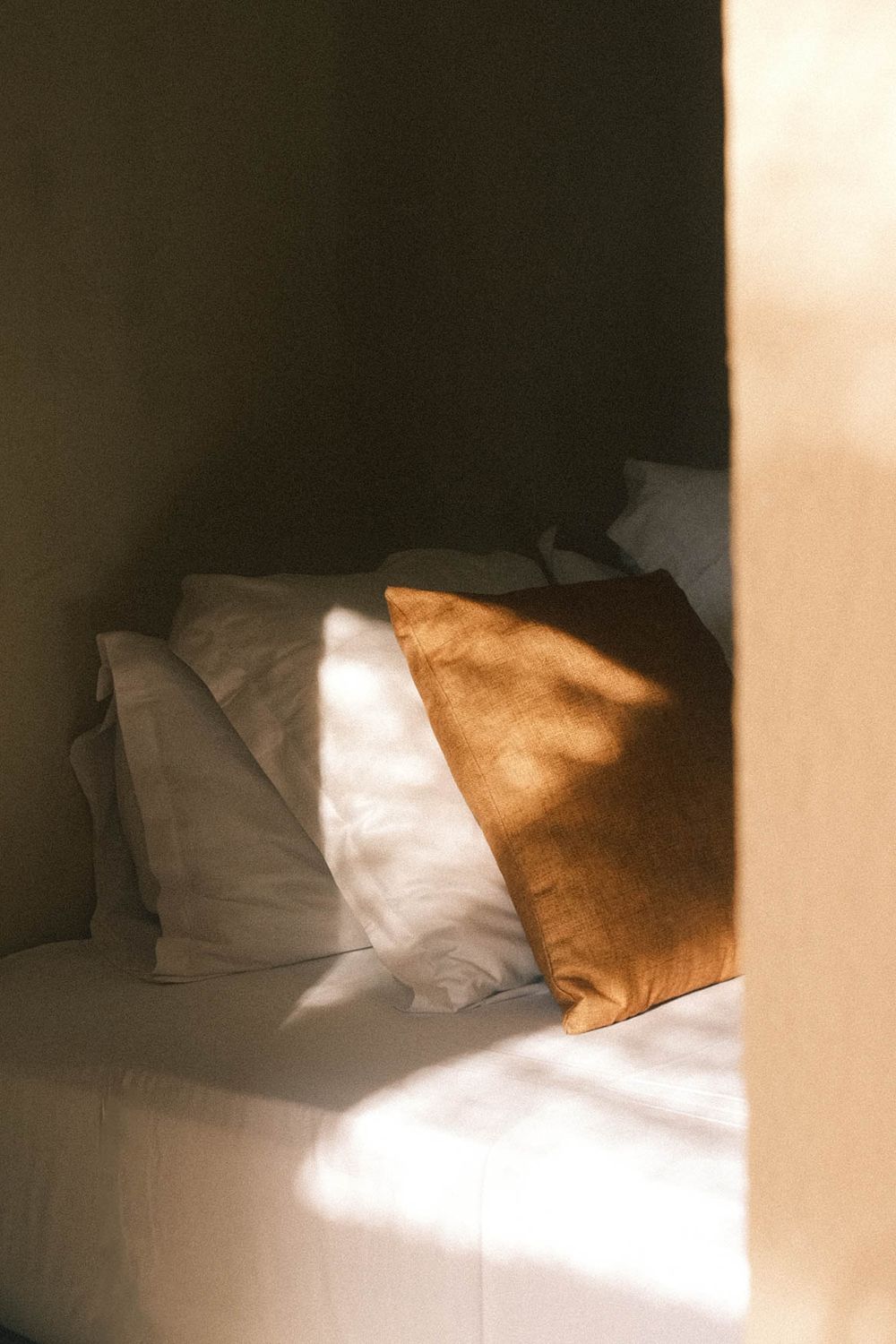 Sunlit bed nook with white linen pillows and a rust-colored cushion casting warm shadows.