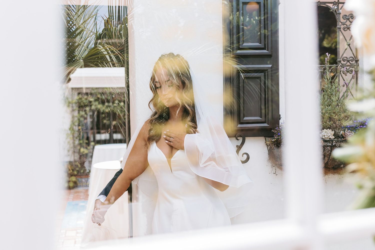 Bride in white wedding dress and veil holding the groom's hand outdoors near elegant dark door and lush greenery.