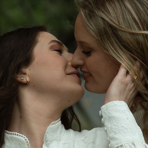 Two women in white dresses share a tender kiss outdoors in a romantic close-up portrait.