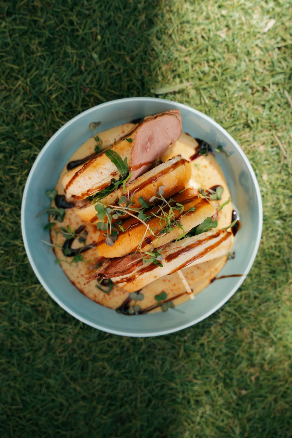 Bol gastronomique avec des tranches de canard, des pêches, des microgreens et un filet balsamique servi sur de l'herbe verte.