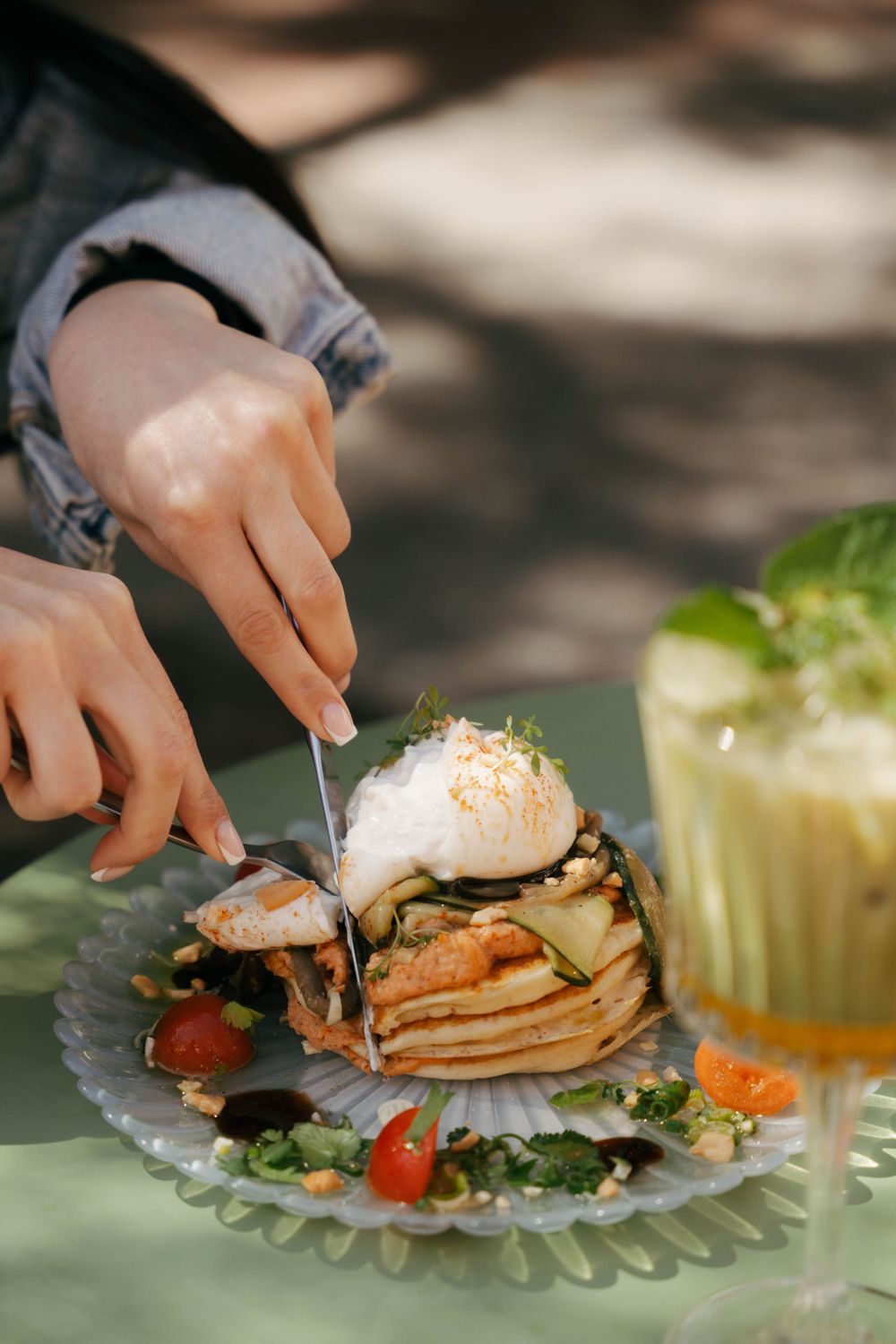 Hands cutting into a layered crepe stack topped with poached egg, served with cherry tomatoes and greens on a glass plate.