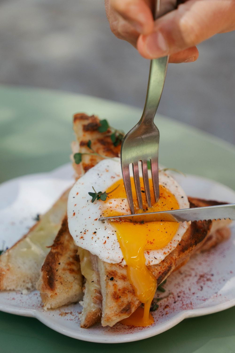 Un œuf poché est ouvert avec une fourchette et un couteau, le jaune coulant sur du pain grillé sur une assiette blanche.