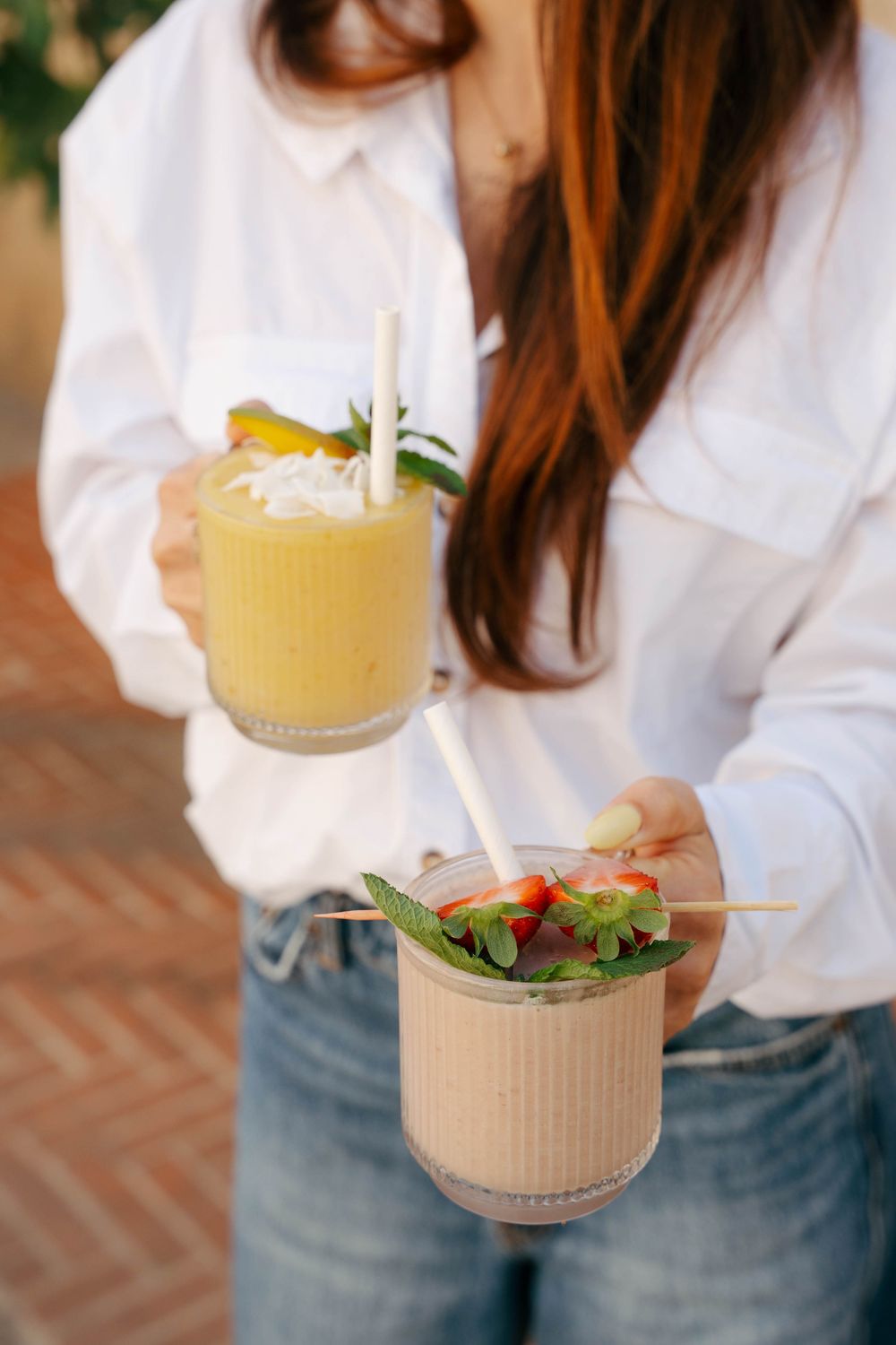 Femme en chemise blanche tenant deux smoothies tropicaux garnis de fruits frais, de menthe et de pailles à l'extérieur.
