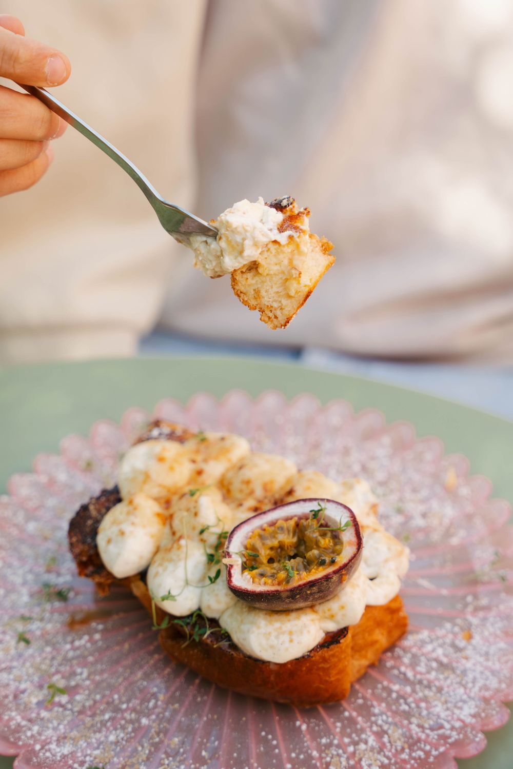 Une fourchette soulevant une bouchée de pain grillé garnie de crème, de fruit de la passion et d'herbes sur une assiette rose.