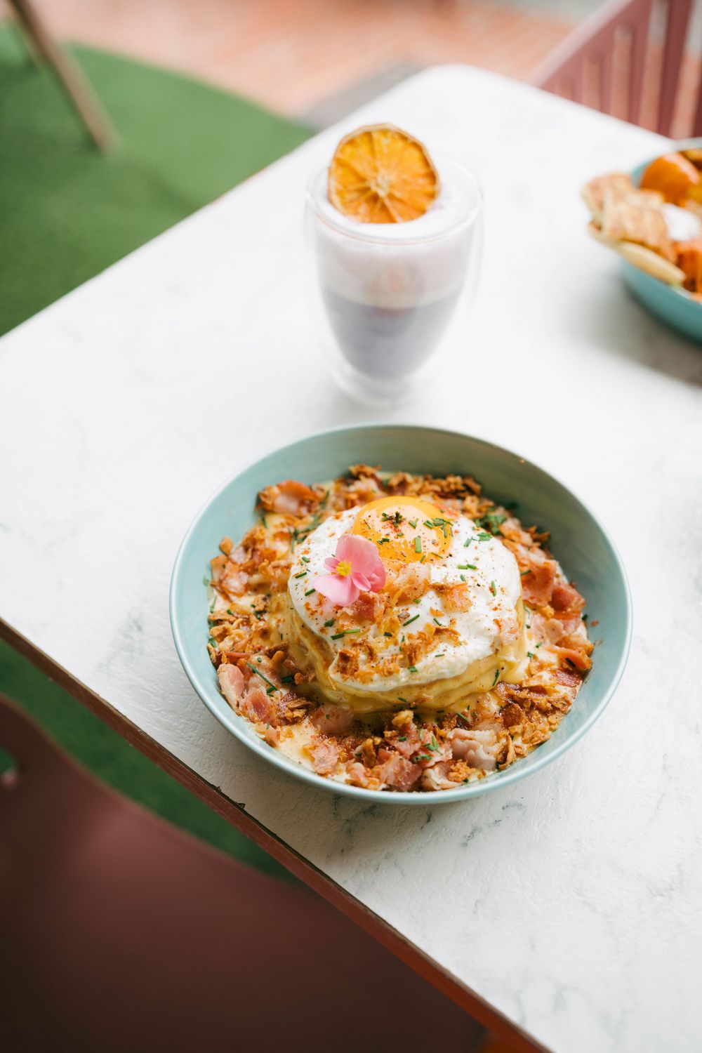 Un bol sarcelle avec des crêpes empilées, un œuf poché, du bacon croustillant et une garniture sur une table en marbre avec une boisson.