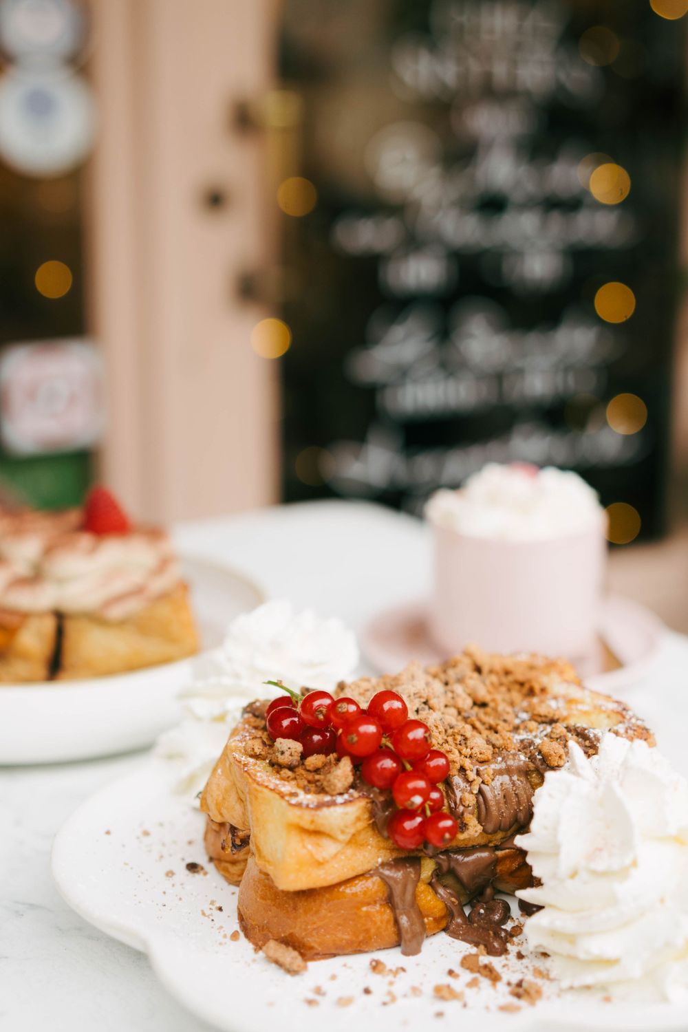 Pain perdu garni de groseilles rouges, de crumble au chocolat et de crème fouettée sur une assiette blanche dans un café confortable.