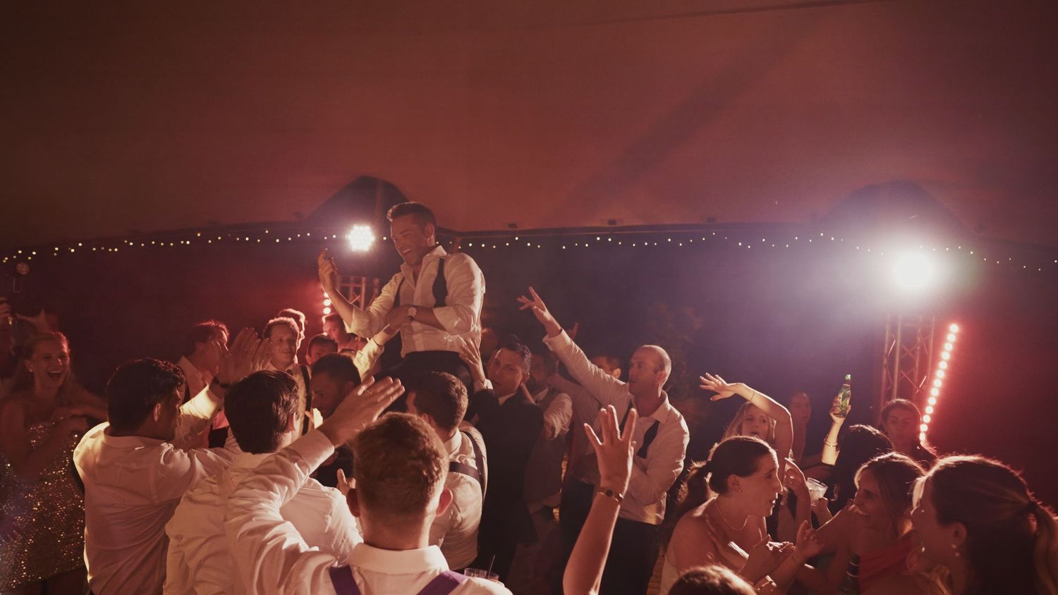 Crowd celebrating at a lively wedding reception with guests cheering and someone lifted up under warm red lights.