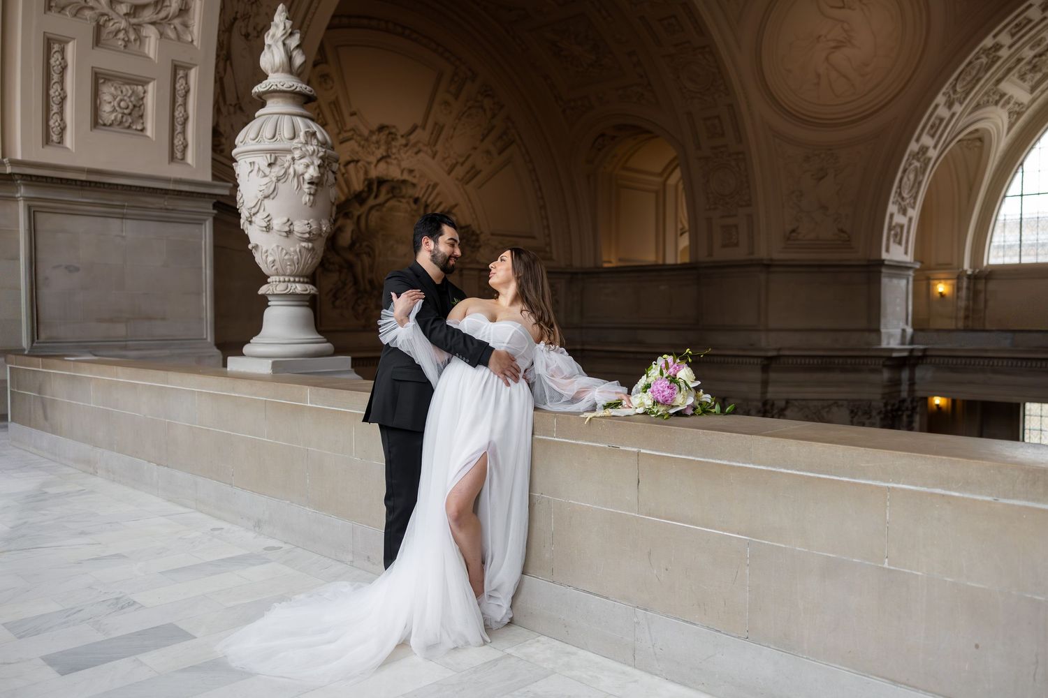 Bride and groom embrace under ornate archway in elegant wedding photo with flowing white dress and formal attire.