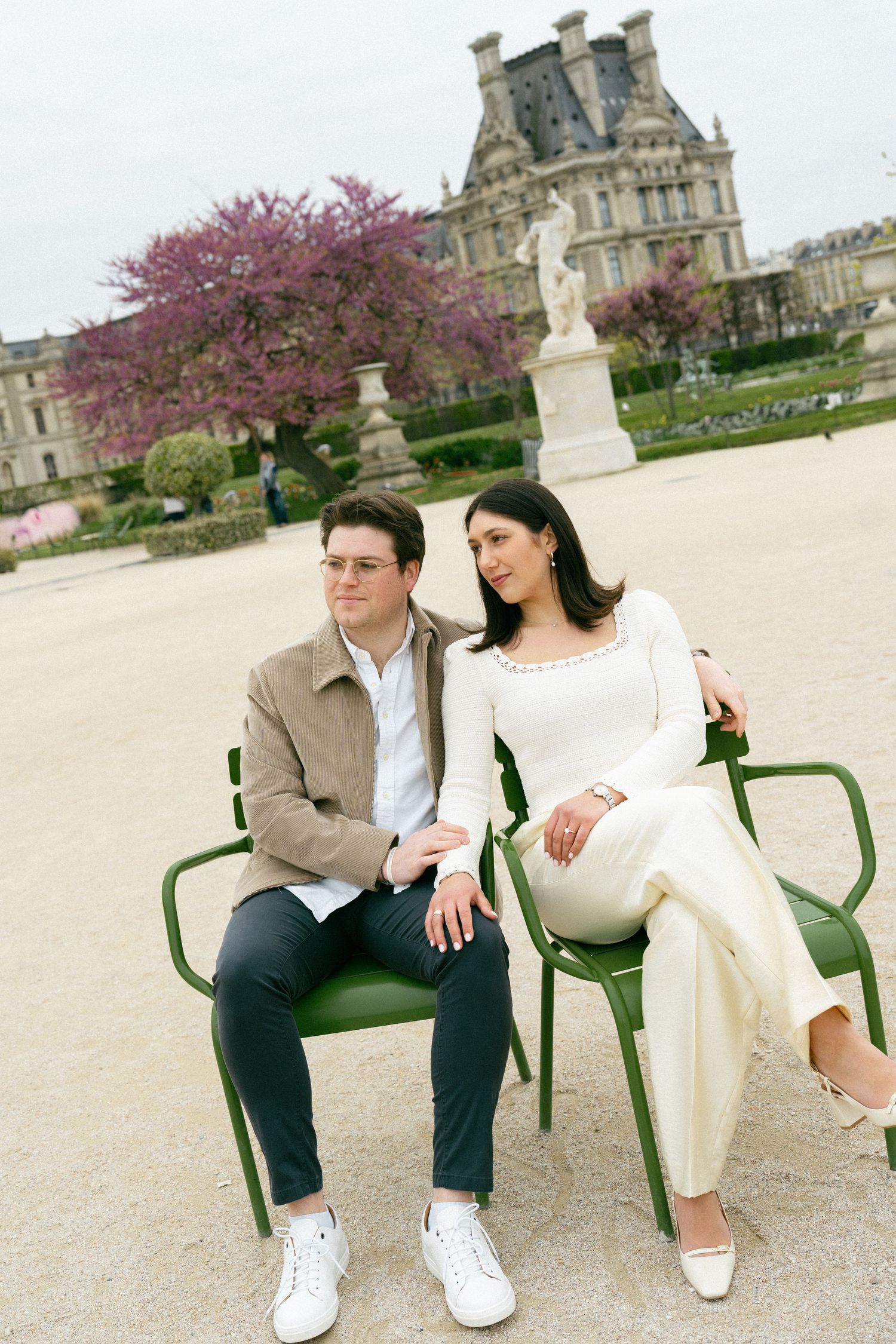 Couple sitting on green chairs in Tuileries Garden, Paris, with blooming cherry trees and the Louvre in the background. Editorial photograph