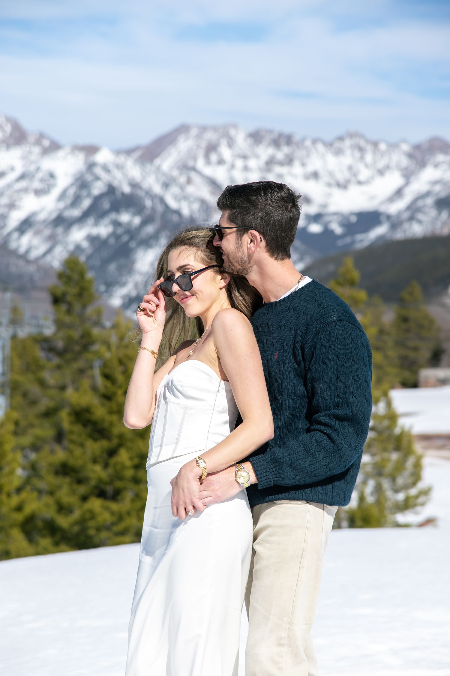Chic cool couple embracing in snow with mountain backdrop, woman in white dress and sunglasses for Vail mountain top engagement.