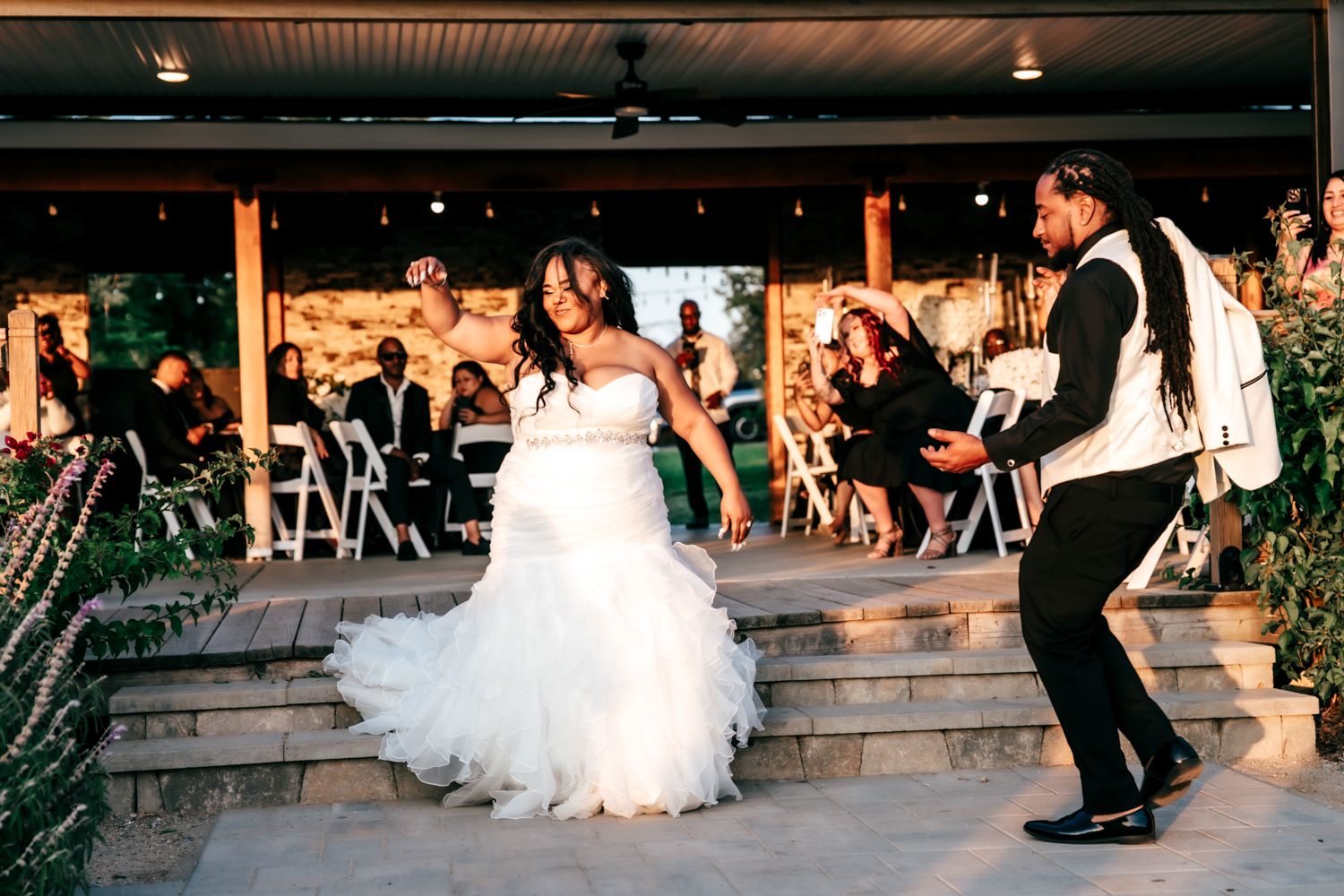 Bride and groom share joyful first dance outdoors at elegant wedding reception with seated guests in background.