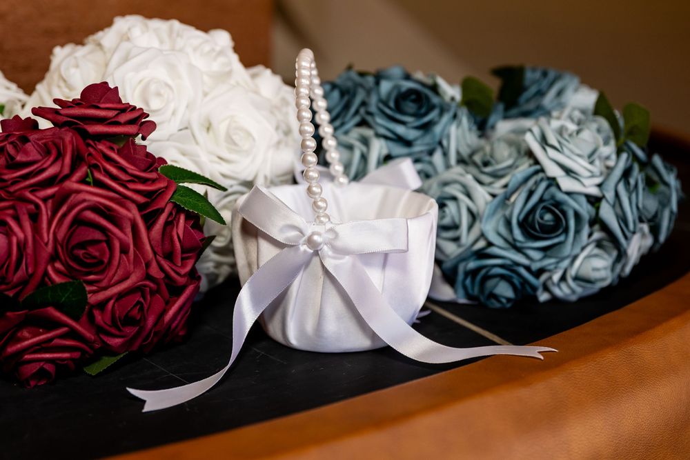 Elegant wedding flower arrangements featuring red, white, and dusty blue roses with a white satin ring bearer basket adorned with pearls.