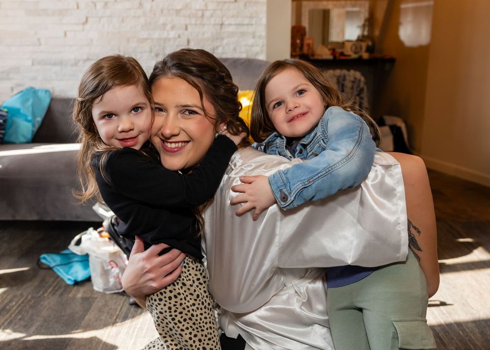 Woman smiling while two young girls hug her from both sides in a cozy living room setting.
