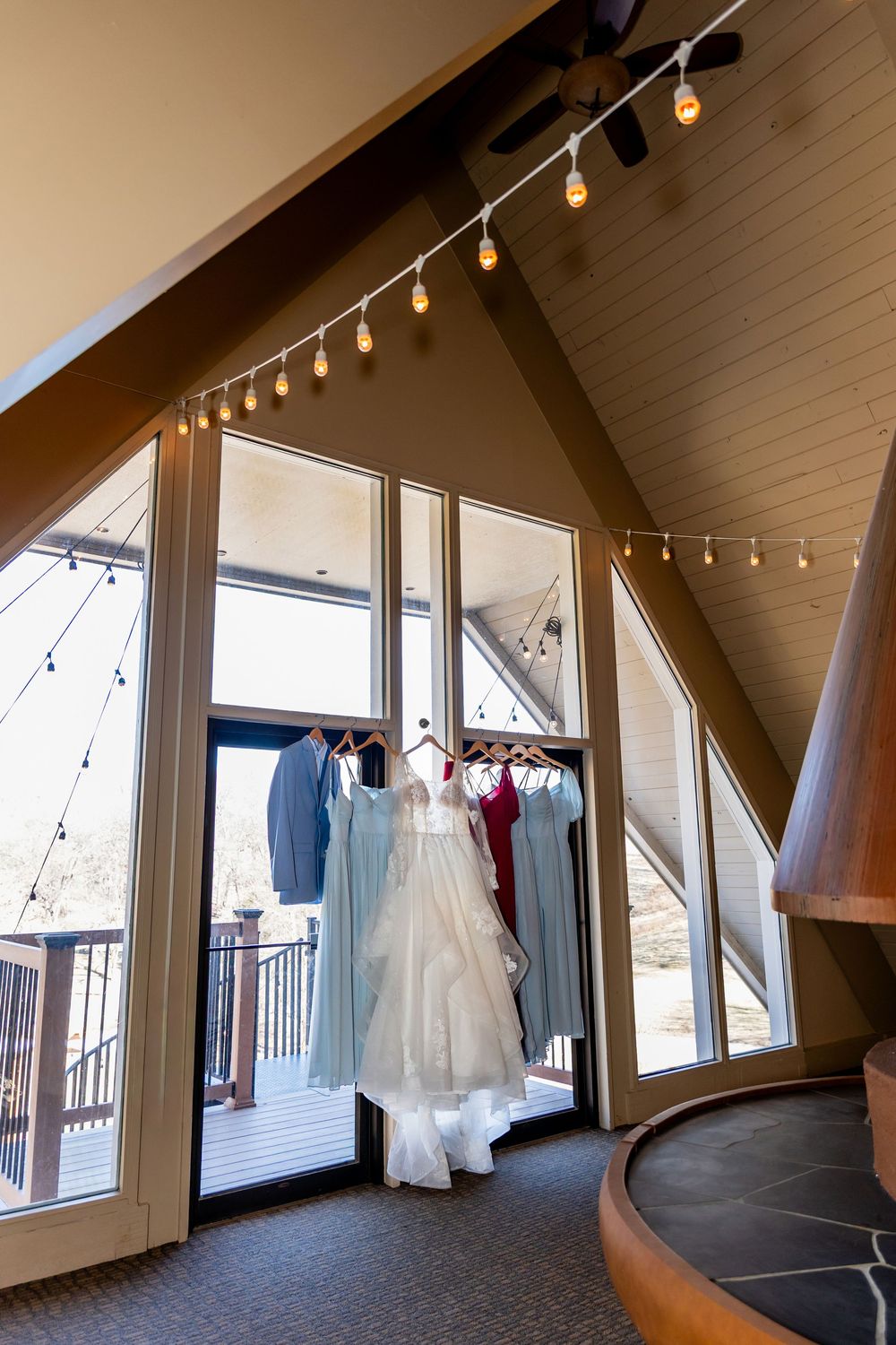 Wedding dress and bridesmaid gowns hanging near a large A-frame window with string lights in an elegant venue.
