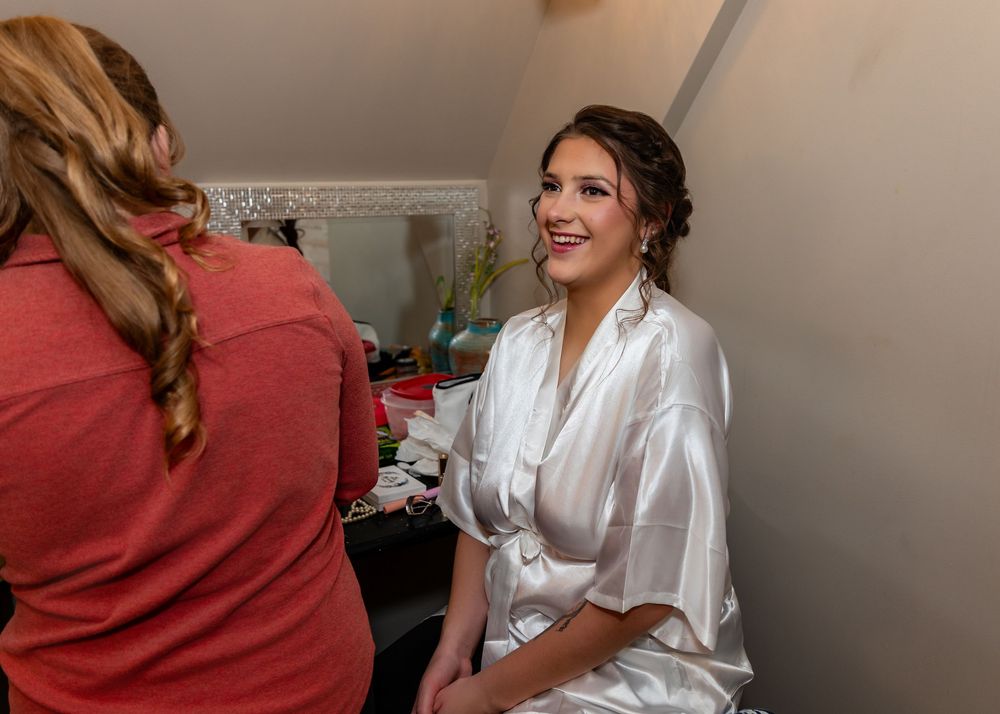 Bride in white satin robe smiling during hair and makeup preparation before her wedding ceremony.
