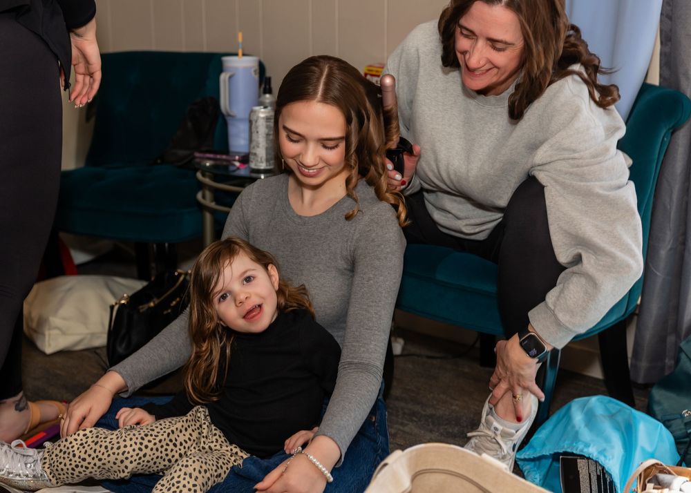 Two women and a young toddler girl smiling and laughing together in a cozy indoor setting.
