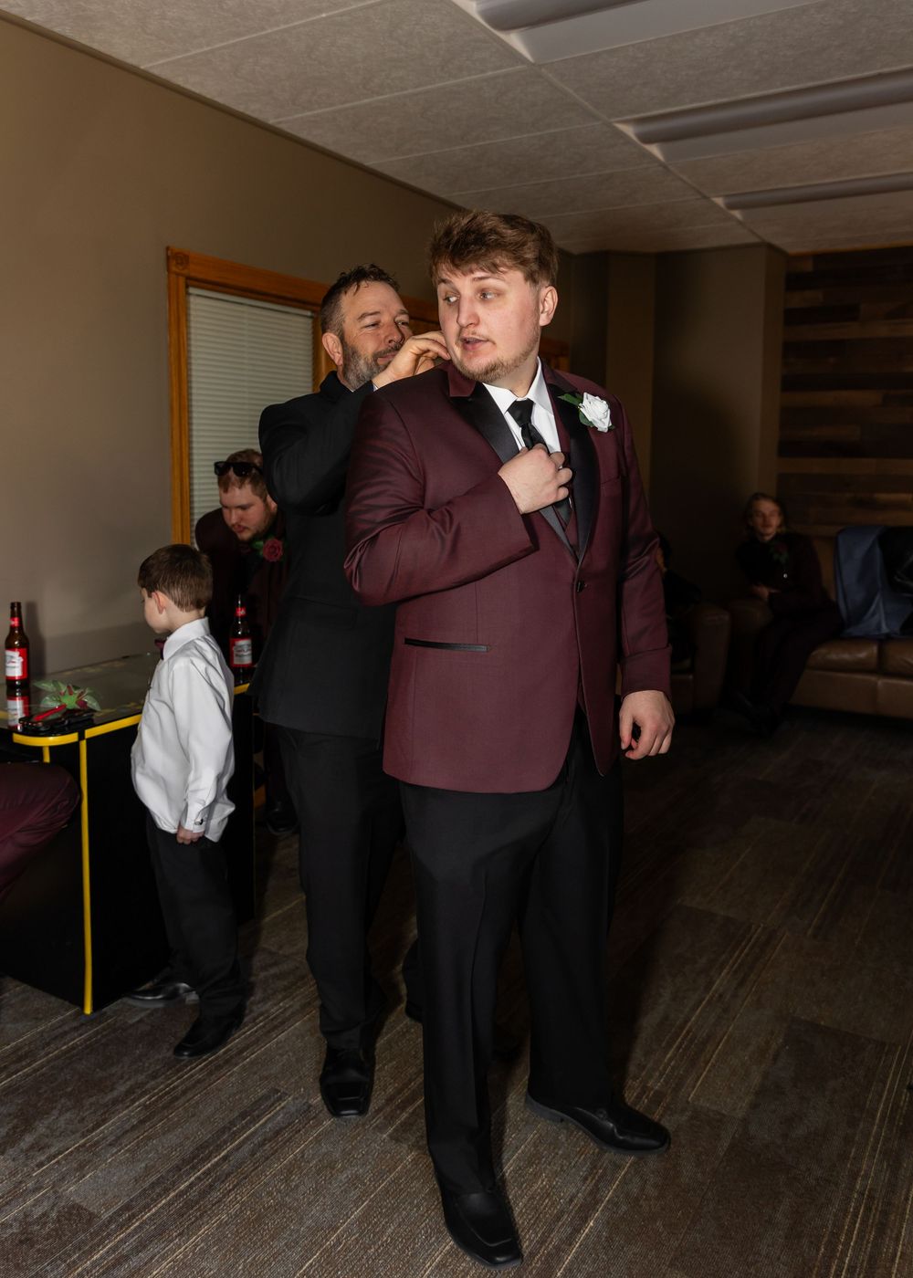 Man in burgundy tuxedo jacket gets tie adjusted by groomsman before wedding ceremony.