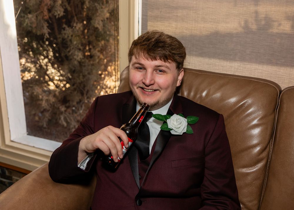 Young man in burgundy tuxedo with white boutonniere sitting in leather chair holding a bottle.