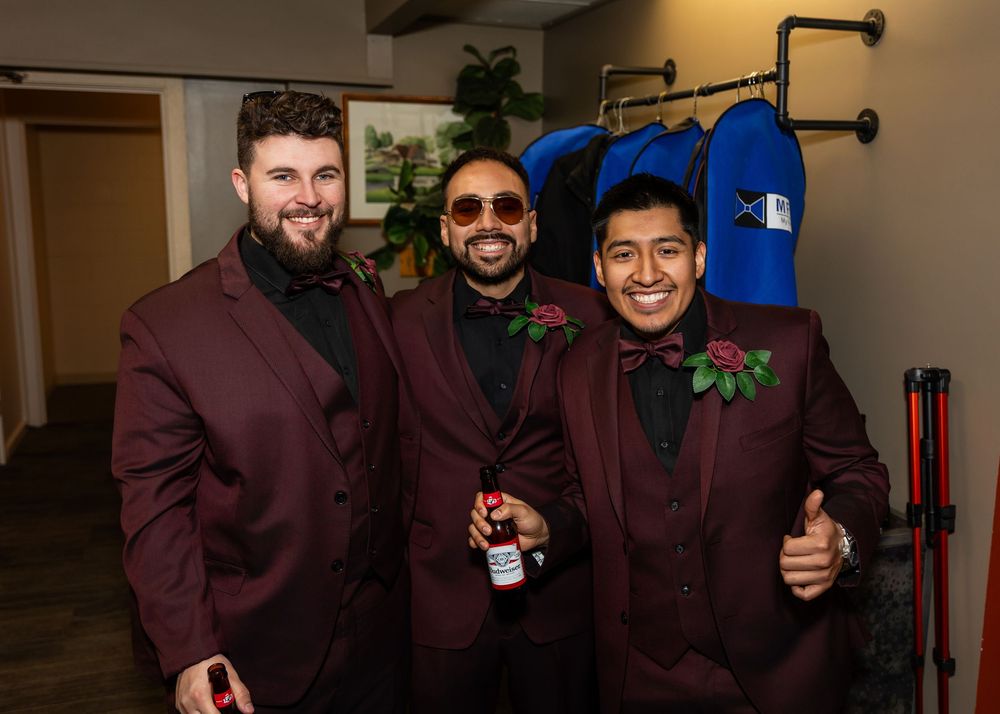 Three men in matching burgundy suits with floral boutonnieres smile together at a formal event.